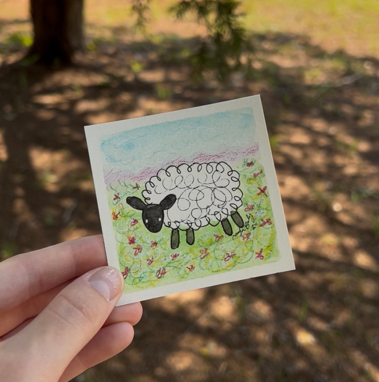 A hand holds a small square art print featuring a hand-drawn black and white sheep in a grassy field under a light blue sky, with a blurred outdoor forest background.