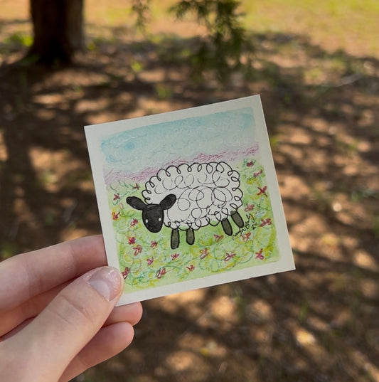 A hand holds a small square art print featuring a hand-drawn black and white sheep in a grassy field under a light blue sky, with a blurred outdoor forest background.